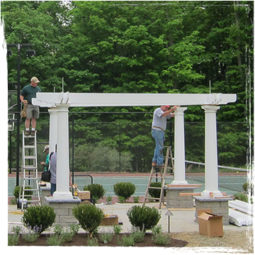 Two men on ladders install beams on a pergola in front of a tennis court
