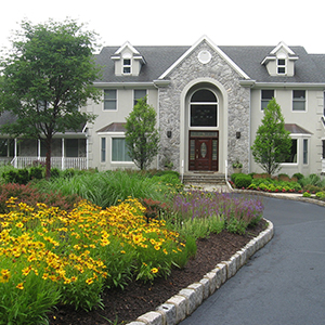 Yellow flowers in a mulched bed lining a paved drive leading to a large grey house